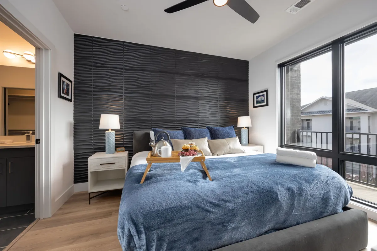 A serene bedroom with a plush bed adorned with a blue blanket, decorative wall accents, nightstands with lamps, and large windows streaming in natural light. The space feels restful and inviting, ideal for relaxation and unwinding after a busy day.