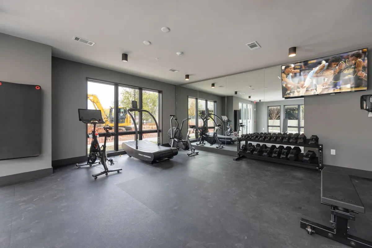 A spacious, well-lit fitness center with treadmills, weight racks, large windows, mirrors, and a television. The atmosphere is motivational, designed for both solo workouts and group fitness classes.