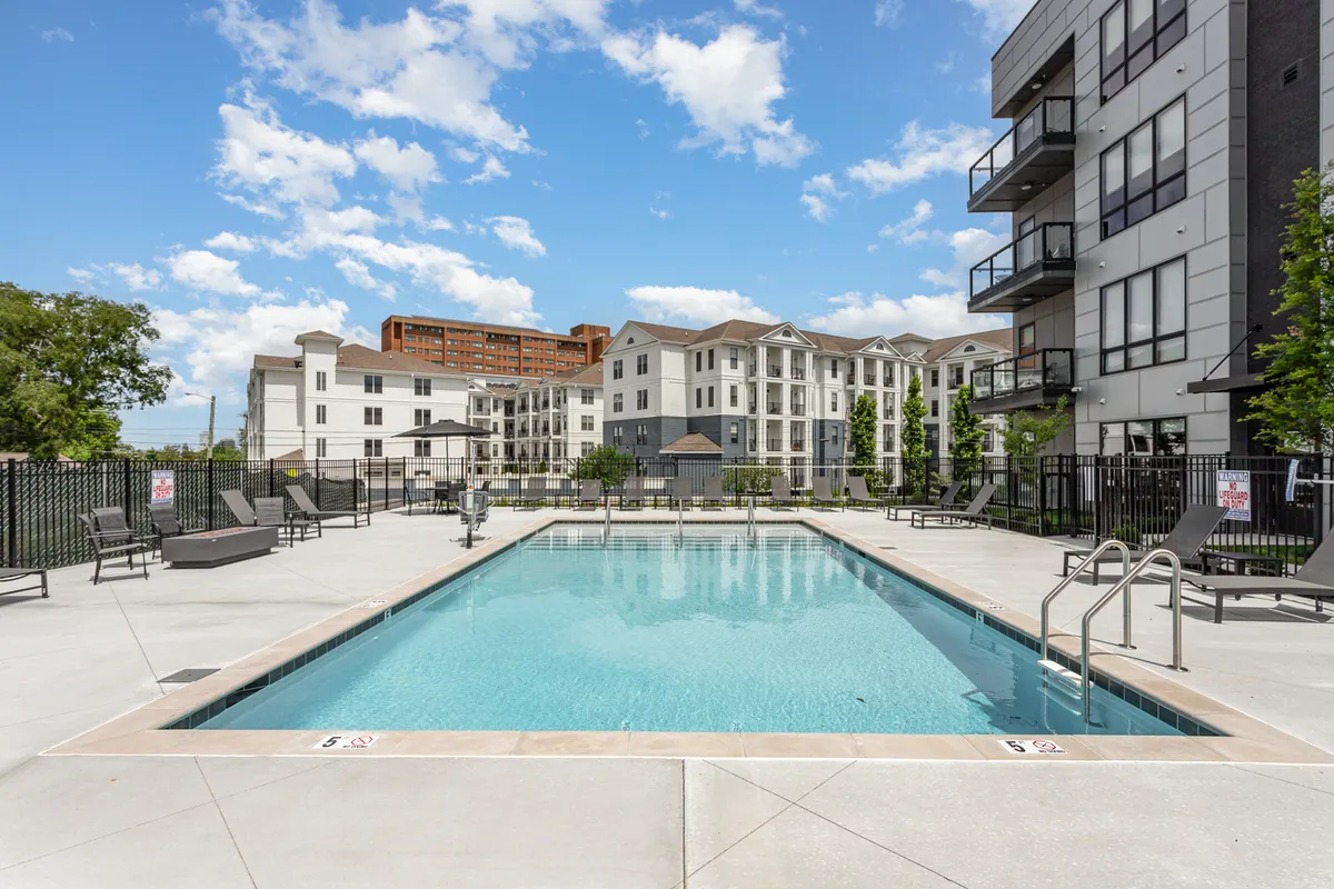 A sun-drenched resort-style swimming pool with lounge chairs arrayed along a landscaped sundeck, surrounded by modern buildings and shaded trees. The inviting blue water and ample seating create an energetic yet relaxed environment ideal for social gatherings.