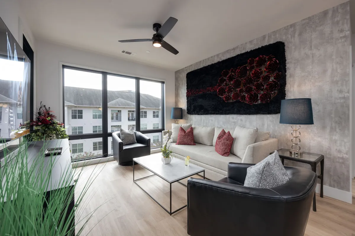 The image captures a bright, inviting living area filled with natural light from a large window. Stylish seating surrounds a modern coffee table adorned with decorative accents, while contemporary artwork adds a touch of flair to the room. The space exudes comfort and sophistication, perfect for relaxing, reading, or socializing.