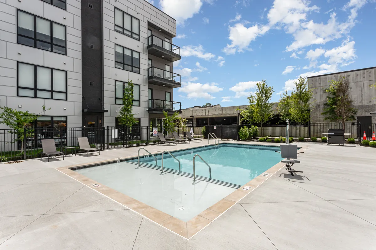 A vibrant exterior view of a swimming pool with balconies overlooking the water, cozy seating areas, and lush greenery. The space appears designed for both relaxation and social events, with visible barbecue and lounge amenities.