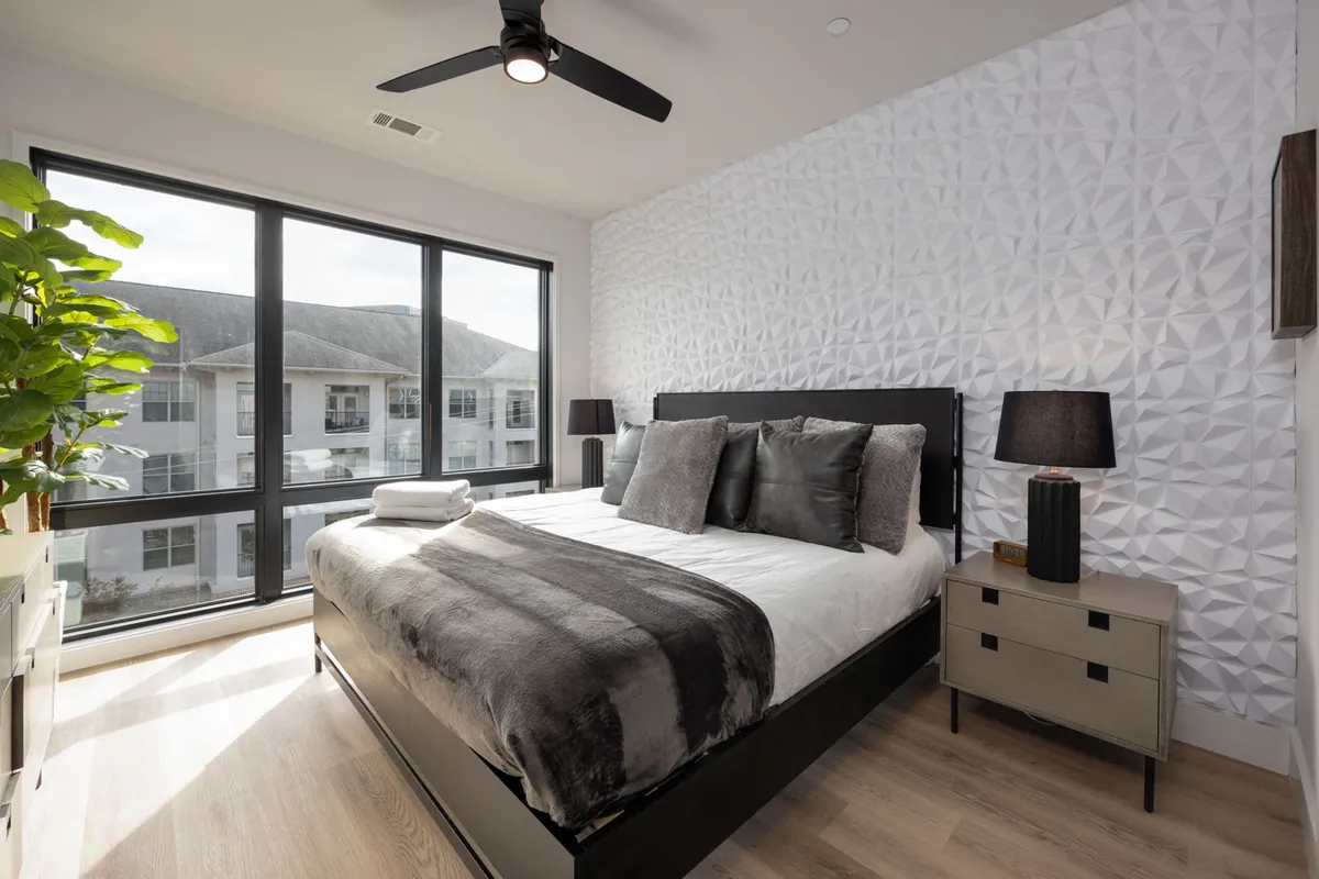 A light-filled bedroom with a plush bed and decorative pillows, a geometric accent wall, large windows offering abundant natural light, an indoor plant, and nightstands with lamps. The decor is contemporary, cozy, and perfect for morning relaxation.