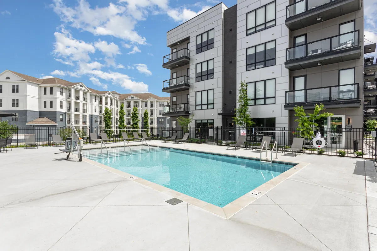 A serene outdoor swimming pool surrounded by a modern apartment complex, with ample landscaped areas and lounge seating. The pool is inviting, suggesting a relaxing and social environment, with hints of sunset lighting enhancing the resort-style vibe.